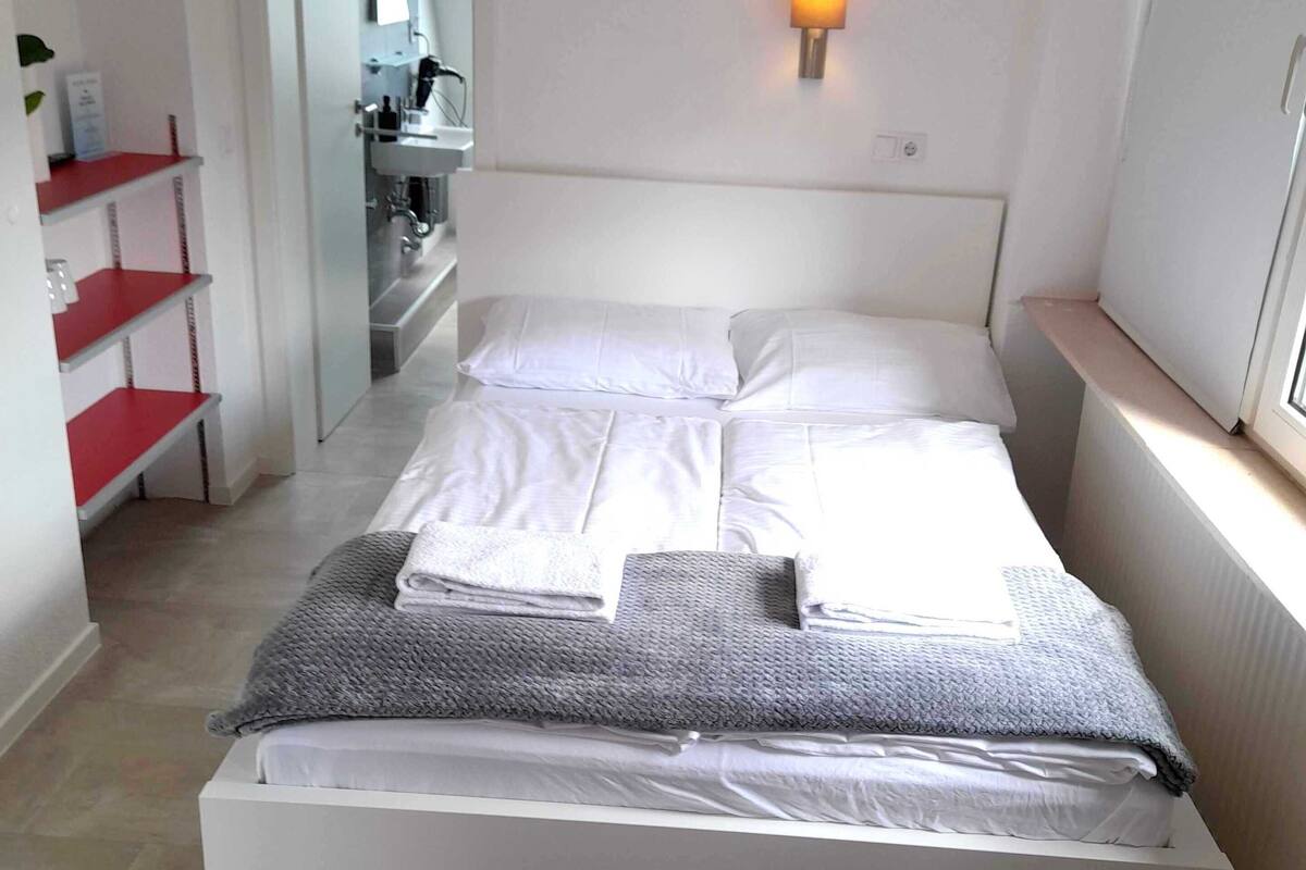 Een slaapkamer met een bed, een nachtkastje, een wandlamp, een raam en een badkamer die zichtbaar is door een open deur.