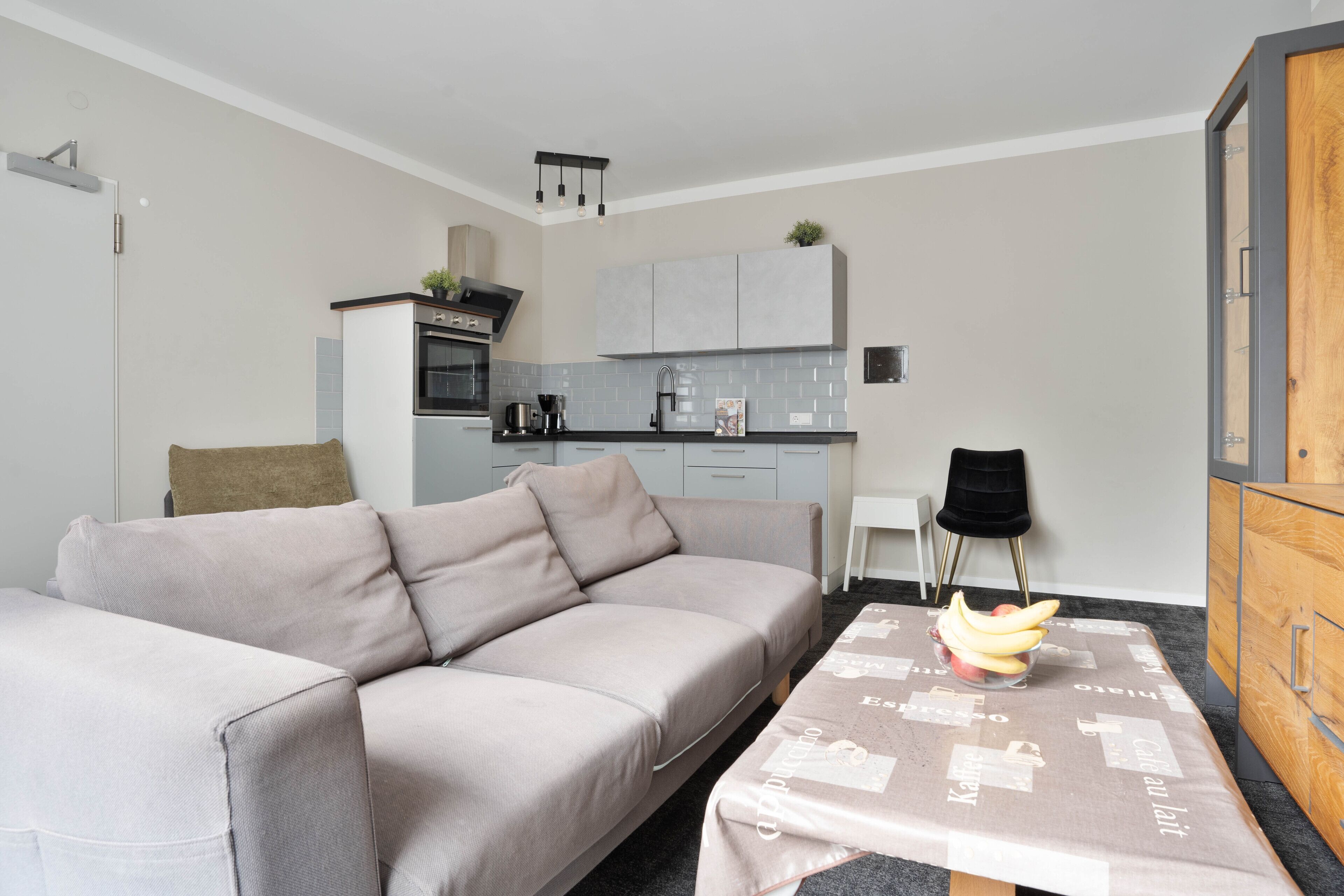 Een moderne woonkamer met een grijze bank, een kleine eettafel met een placemat, een keuken op de achtergrond en een houten kast.