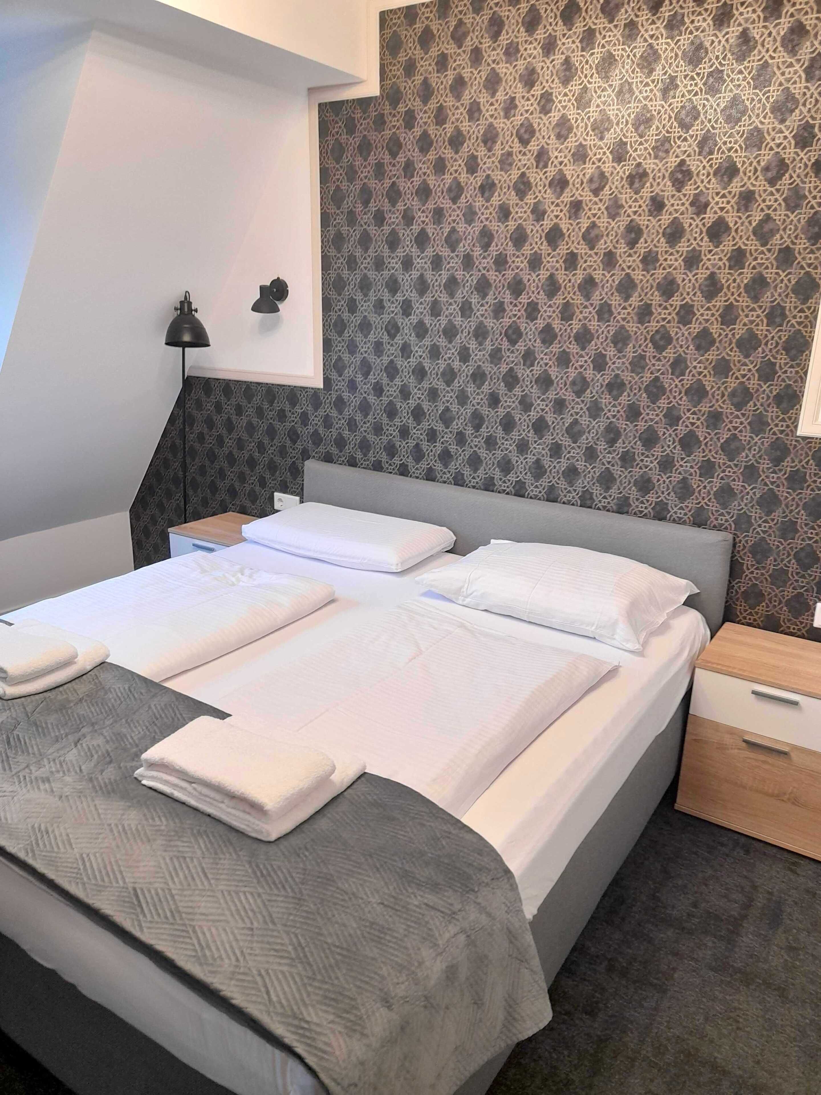 A modern hotel room with a large bed, a bedside table, a nightstand, and a patterned wallpaper.