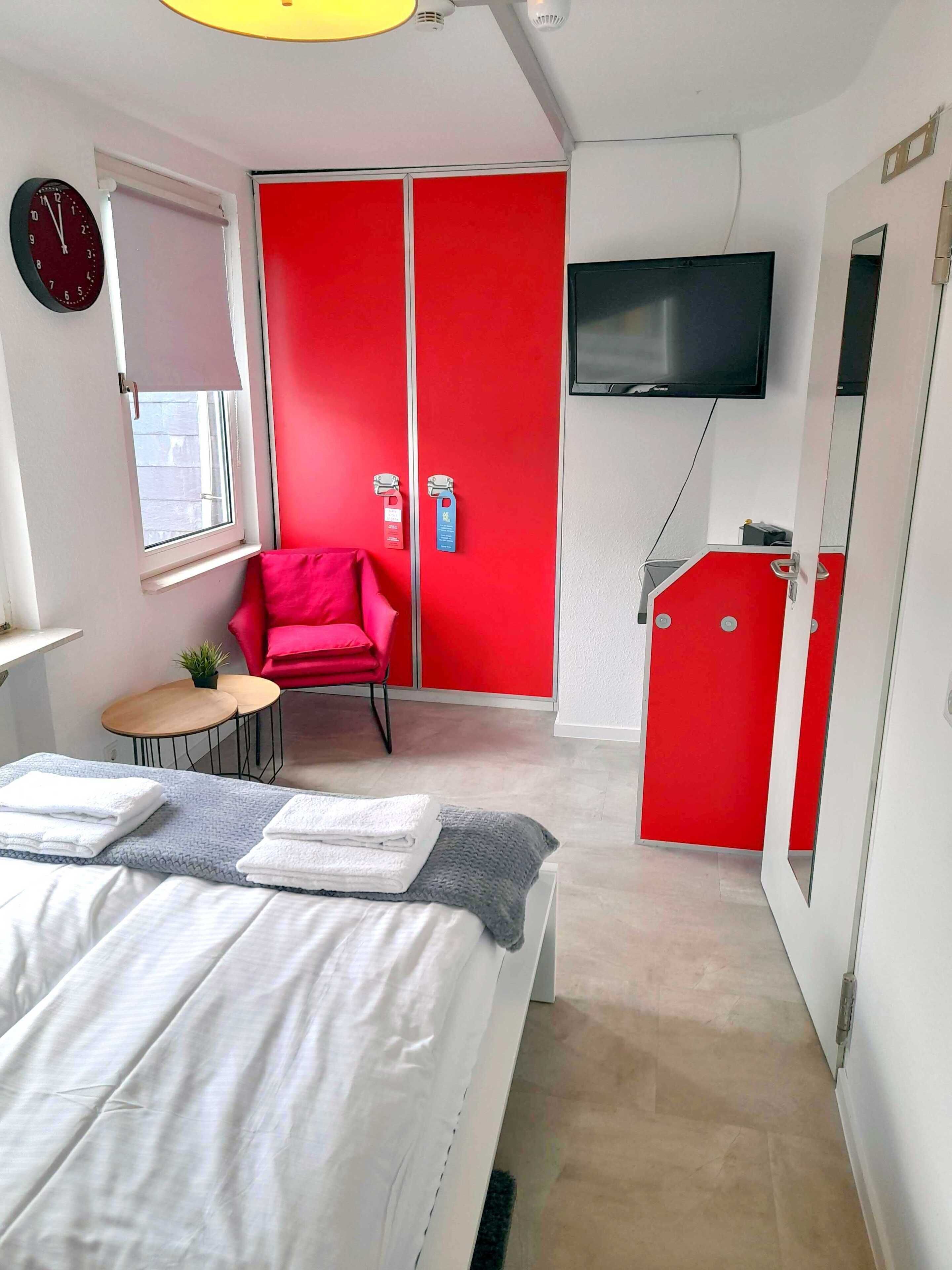 Een hotelkamer met een bed, een rode garderobe, een televisie, een rode stoel en een klein tafeltje met een plant.