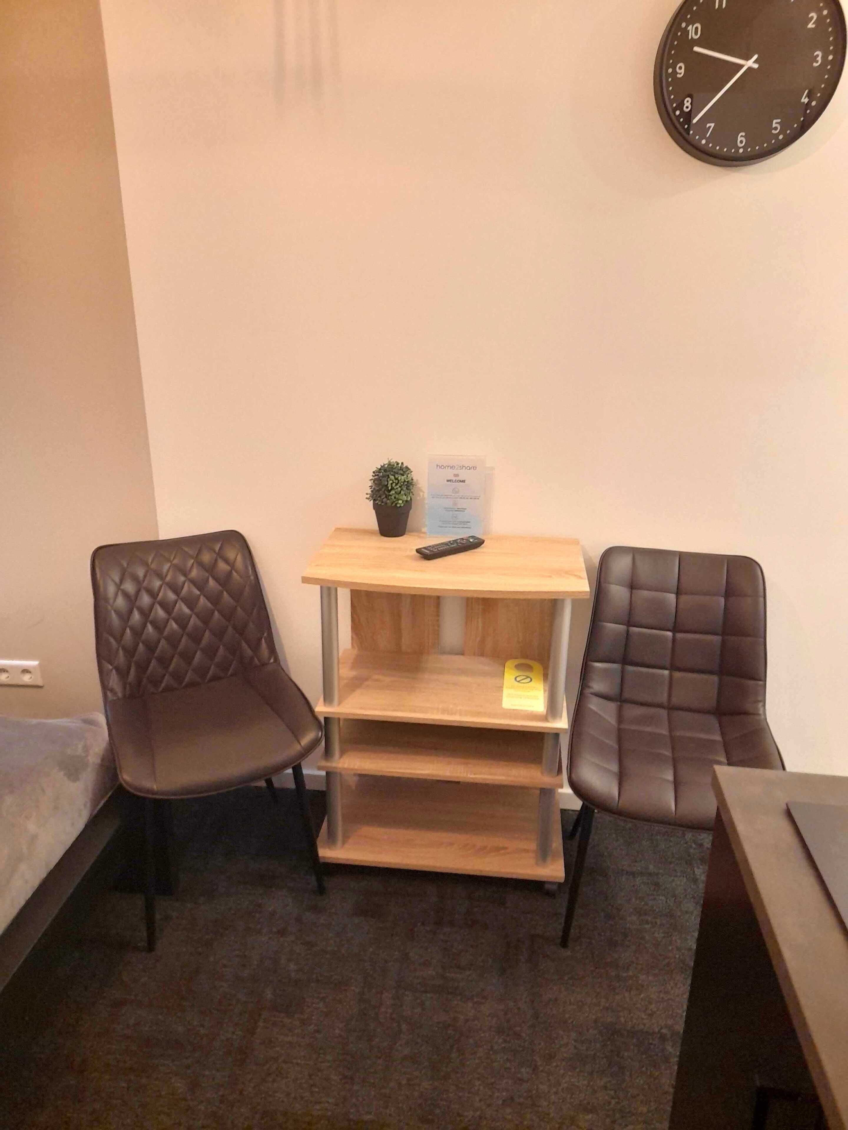 Una habitación pequeña con dos sillones de cuero marrón, un escritorio de madera con una planta y un control remoto, y un reloj de pared.