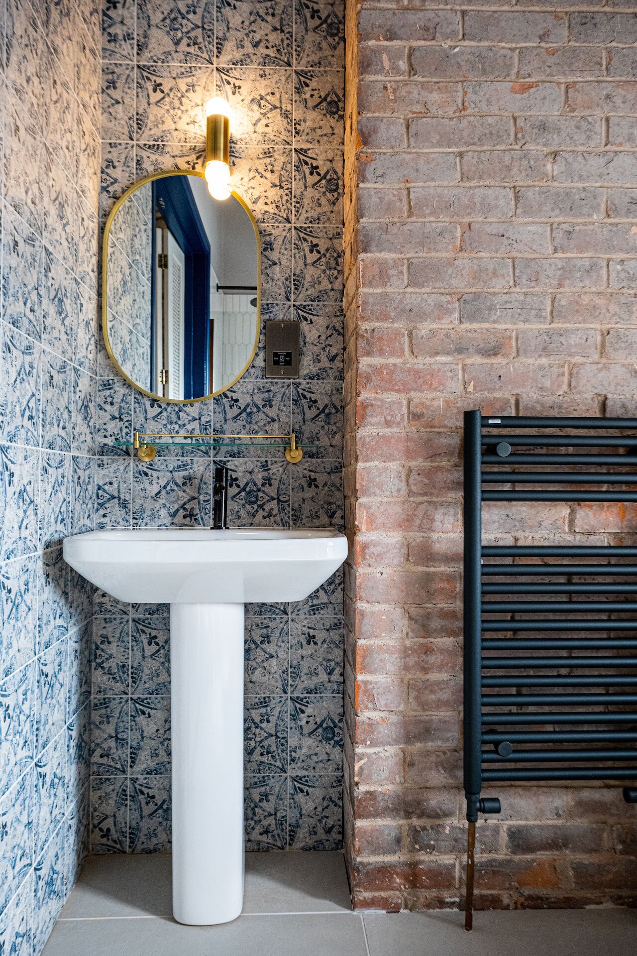 A bathroom with a white pedestal sink, a round mirror with a gold frame, and a black radiator.