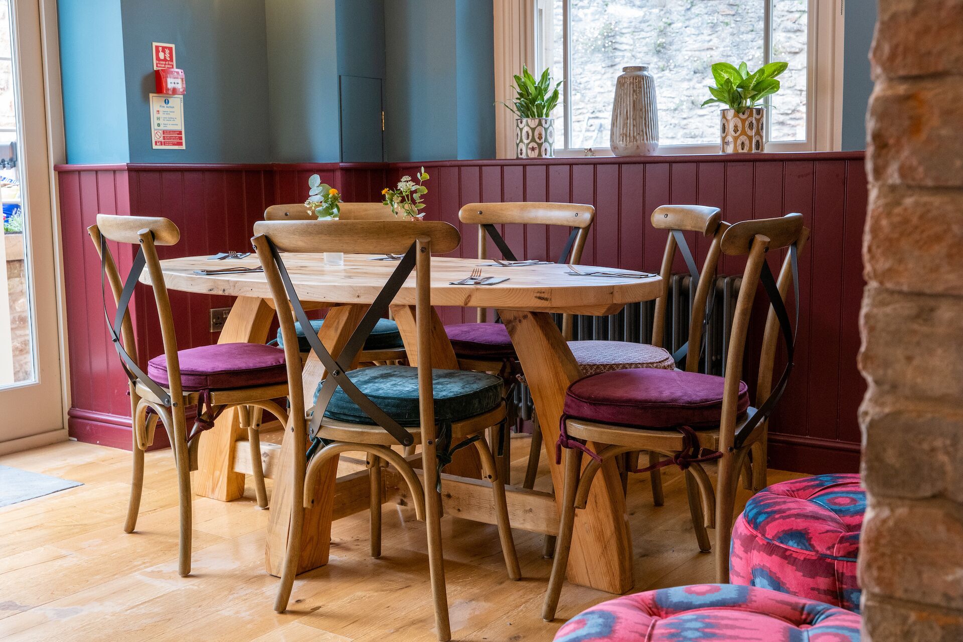 A dining area with a round wooden table surrounded by six wooden chairs with cushioned seats. The room has wooden flooring and a window with a view of the outside.