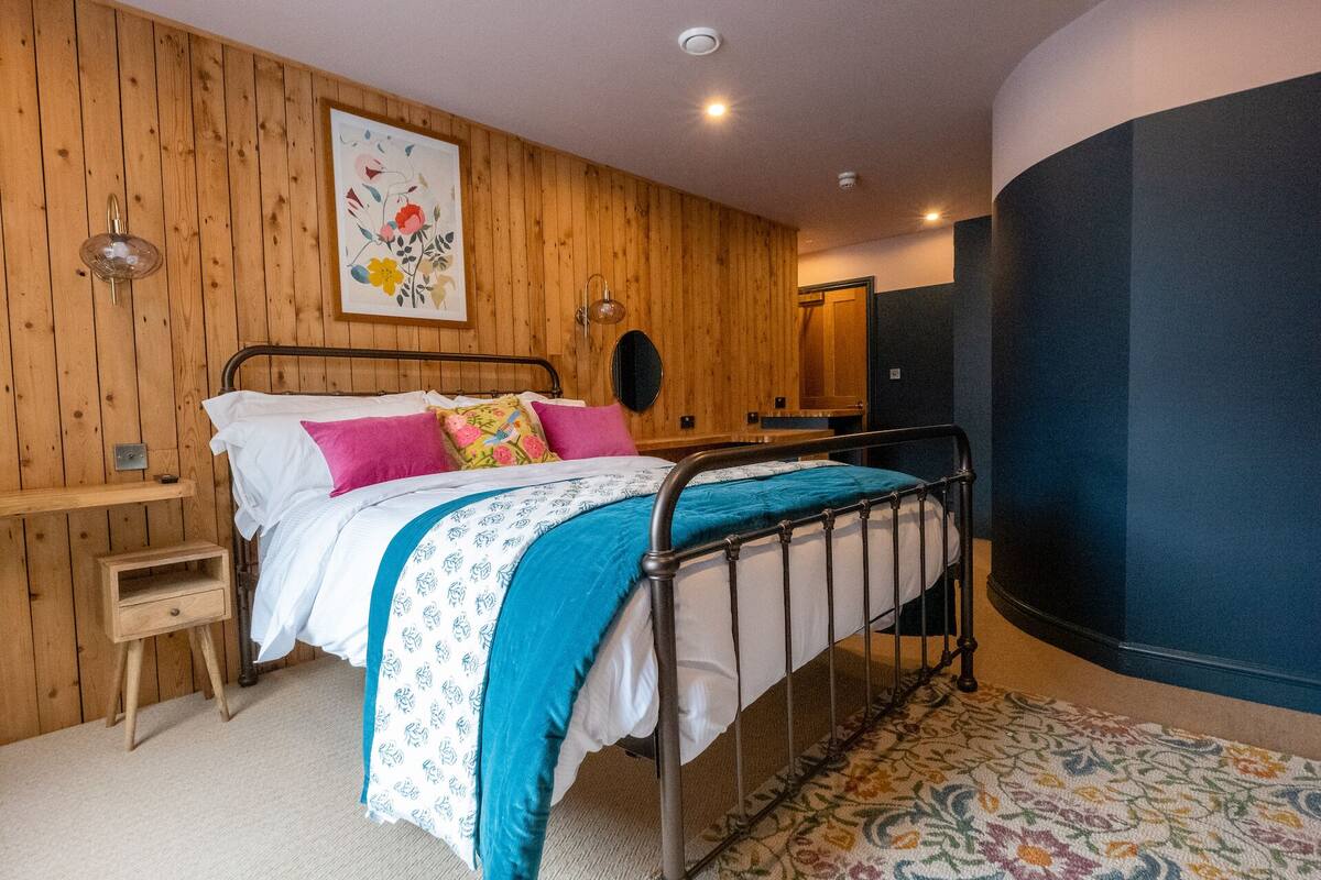 A bedroom with a wooden wall, a bed with a metal frame, a nightstand, a mirror, and a floral-patterned rug.