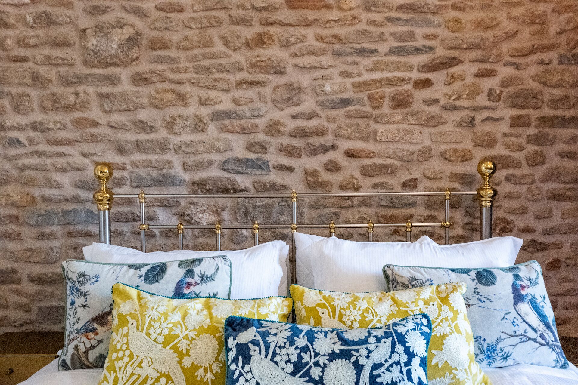 Una cama con estructura metálica y detalles dorados ornamentales, frente a una pared de piedra.