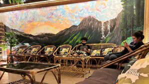 A room with wicker furniture, a large mural of mountains, and a Forrest Hostels pillow. - Forrest Hostels (Mumbai)
