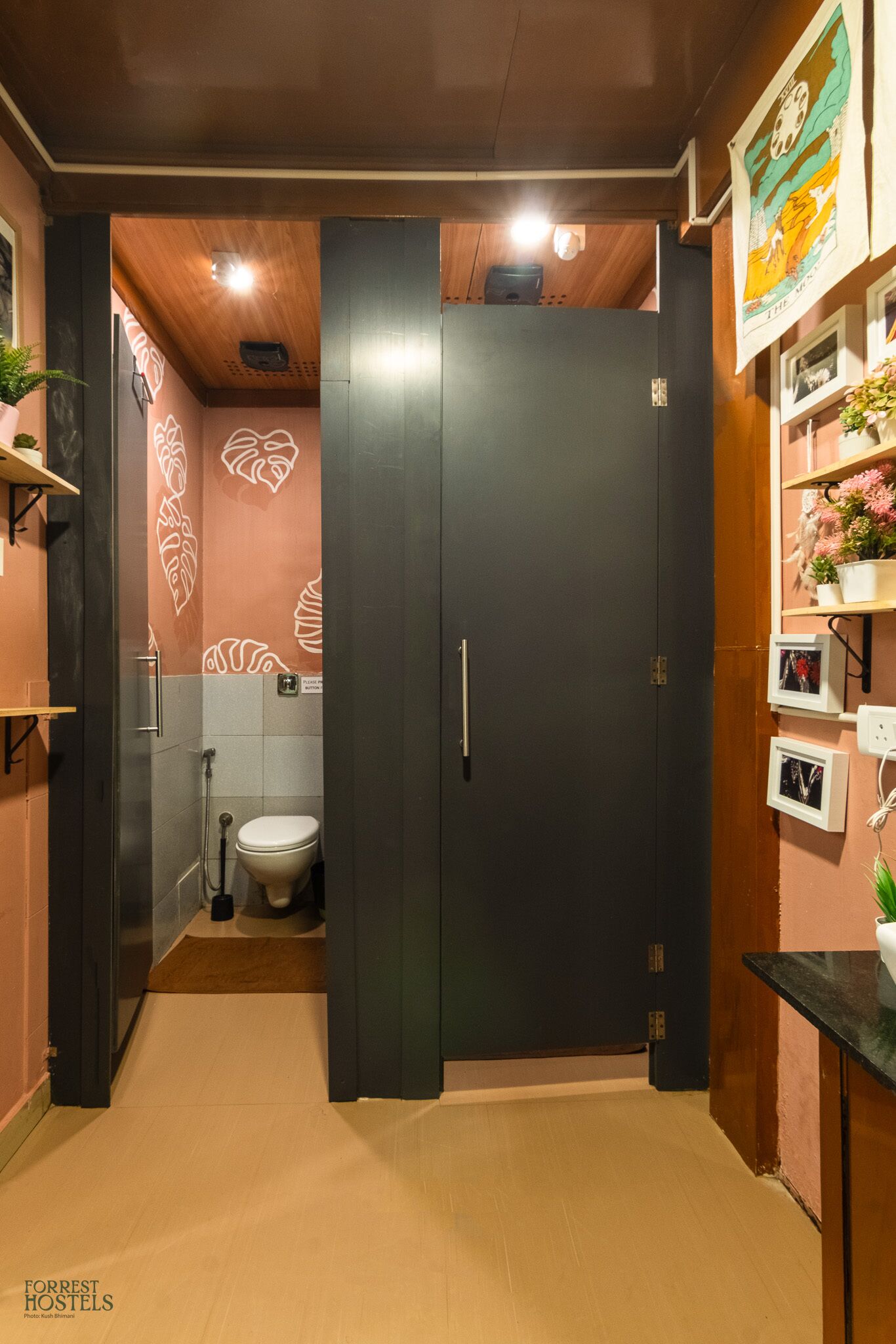 A bathroom with a toilet, shower, and a dark door. There are shelves with plants and a wall with a floral pattern.