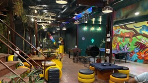 A vibrant indoor space with a green wall mural, a ping pong table, and a variety of seating options including yellow and black stools. - Forrest Hostels (Mumbai)