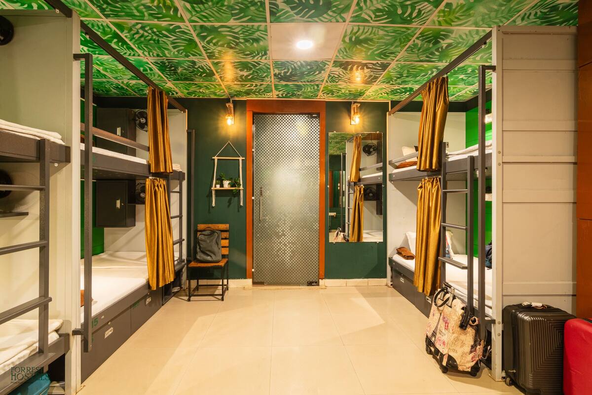A dormitory room with bunk beds, a small desk, a chair, a mirror, a wardrobe, and a suitcase.