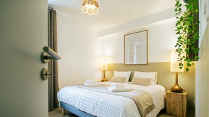Une chambre à coucher bien rangée, avec un lit, des lampes de chevet, une œuvre d’art accrochée au mur et une plante en pot.