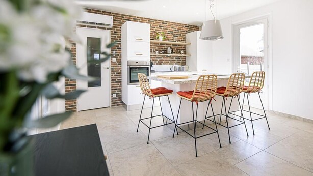 Une cuisine moderne avec un mur recouvert de briques, des meubles blancs et un coin repas agrémenté de chaises et d’une table en osier.