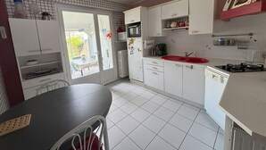 A compact kitchen with white cabinets, a red sink, and a dining area with a table and chairs. - Charming 2-Bedroom House with Garden Near Beach and Port Olona (Les Sables-d'Olonne)