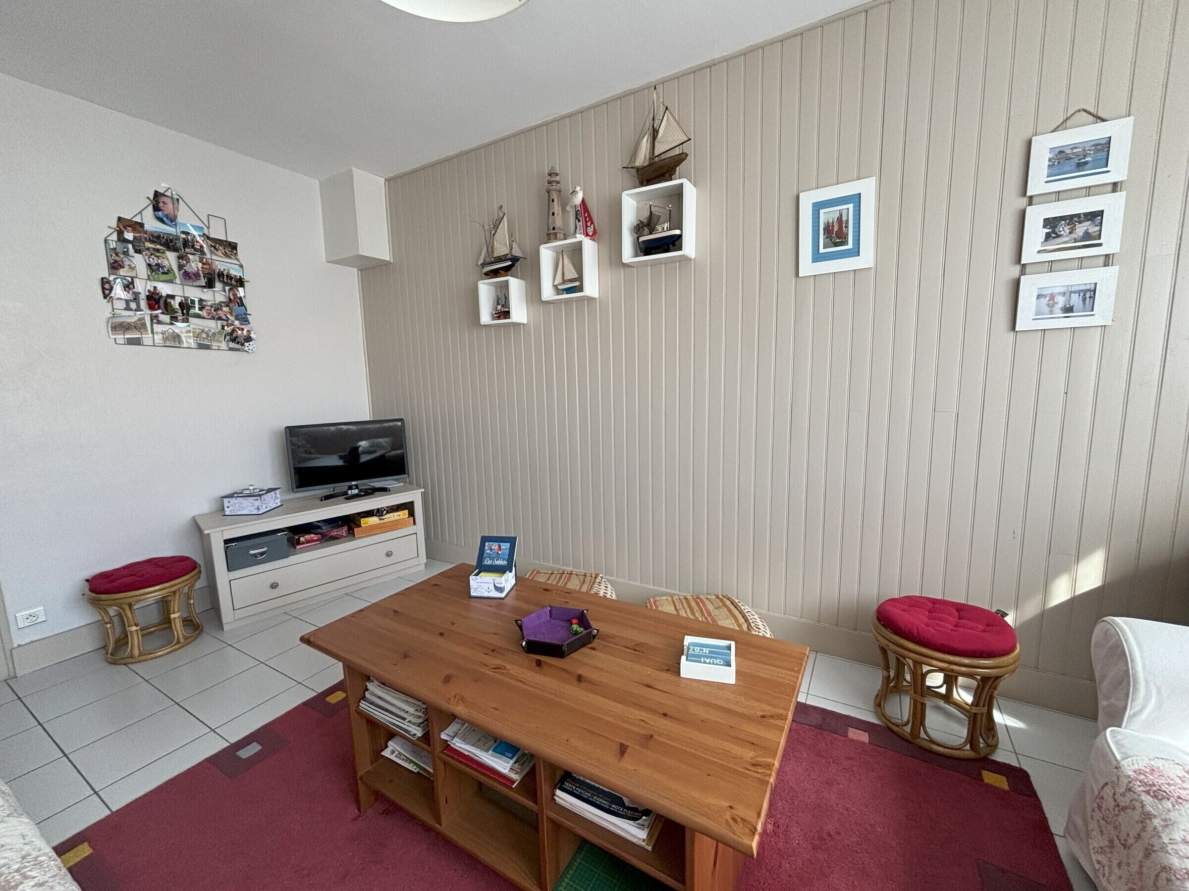 Ein gemütliches Wohnzimmer mit einem Holztisch, einem Fernseher auf einer weißen Kommode und einer Wand, die mit gerahmten Fotos und kleinen Schiffmodellen dekoriert ist.
