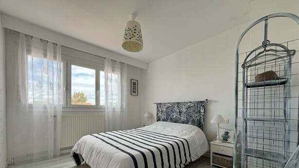 A bedroom with a bed, a nightstand, a hanging light fixture, a window with curtains, and a metal rack. - Charming 2-Bedroom House with Garden Near Beach and Port Olona (Les Sables-d'Olonne)