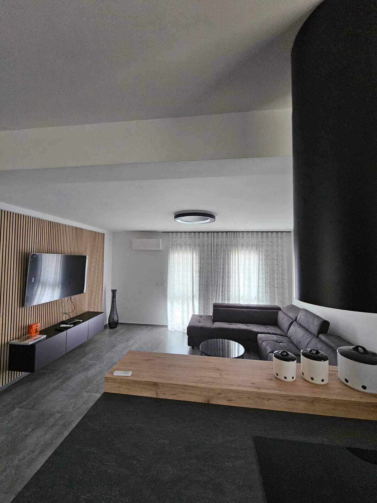 A modern living room with a sectional sofa, a flat-screen TV mounted on a wooden panel, a round coffee table, and a large black accent wall.