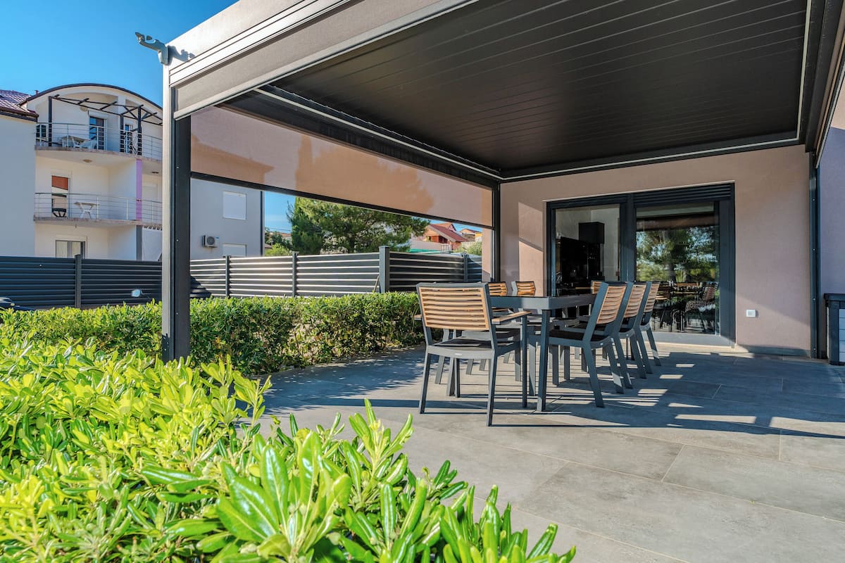 A covered outdoor seating area with a table and chairs, surrounded by greenery.