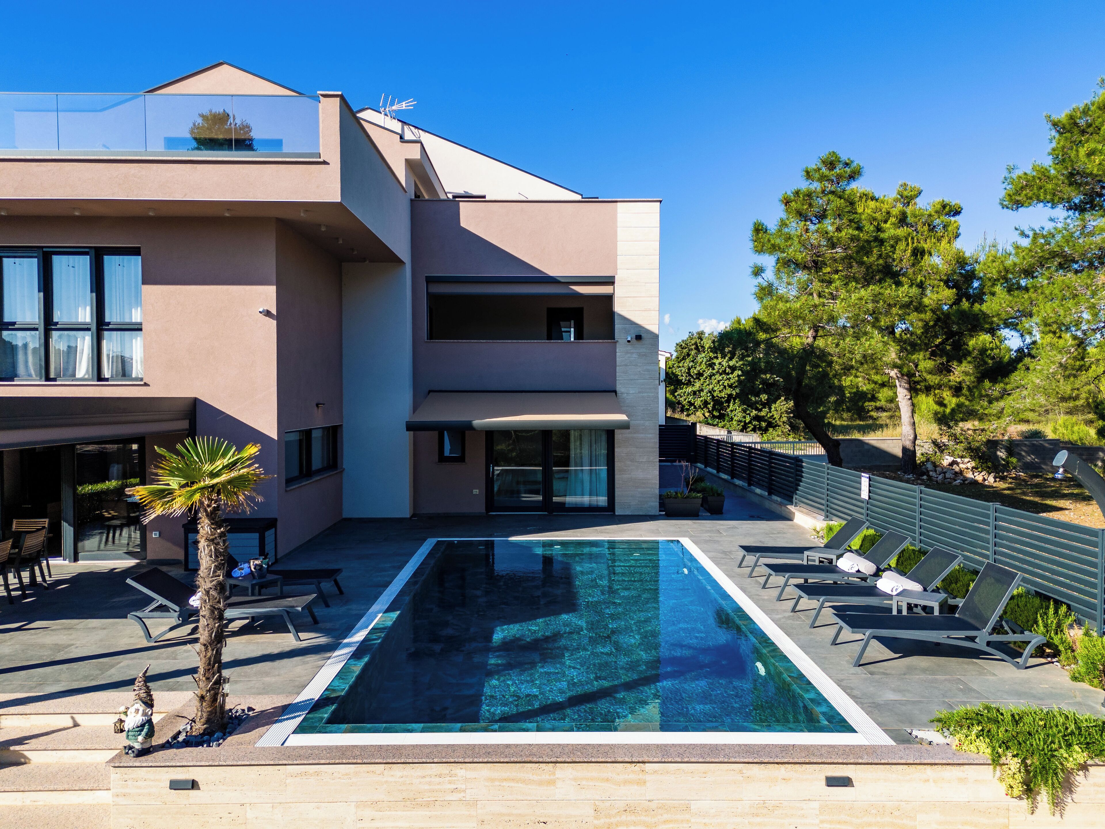 Et moderne hus med to etager, en swimmingpool, udendørs møbler og udsigt til træer.