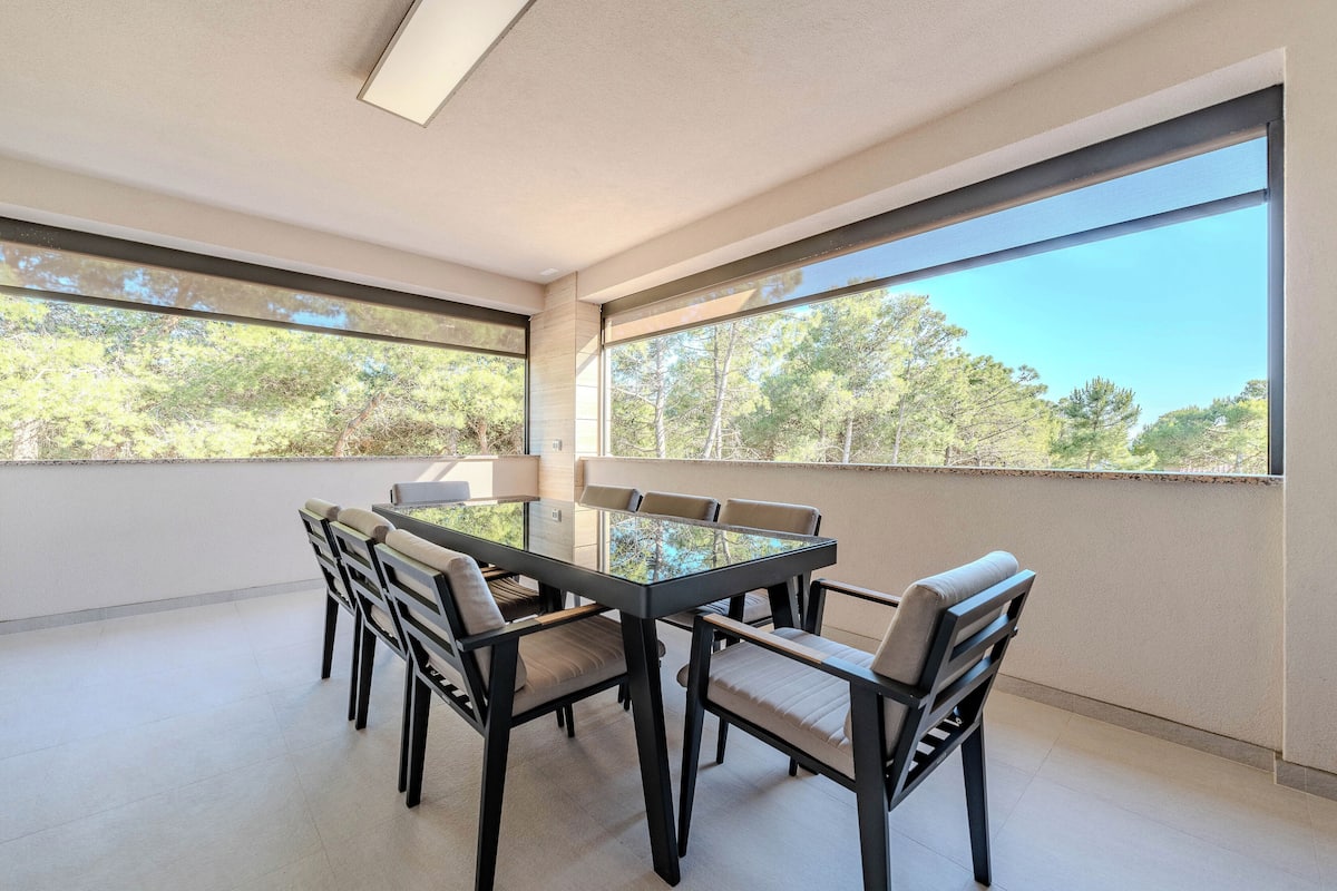 A modern dining area with a glass table and chairs, large windows offering a view of trees.