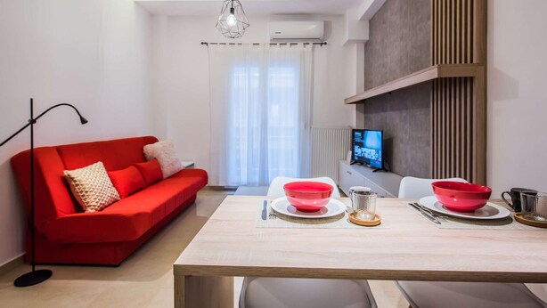 Een moderne woonkamer met een rode bank, een eettafel met rode kommen, een tv op een houten plank en een groot raam met dunne gordijnen.