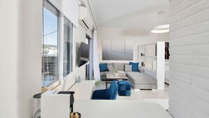 A modern living room with a sofa, ottomans, and a coffee table. There is a TV mounted on the wall and a window with blinds. - Pearl: Penthouse suite with city view (Thessaloniki)