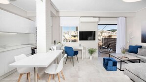 A modern apartment with a kitchen, dining area, and living room. It features a large window with a city view, a balcony, and a flat-screen TV mounted on the wall. - Pearl: Penthouse suite with city view (Thessaloniki)