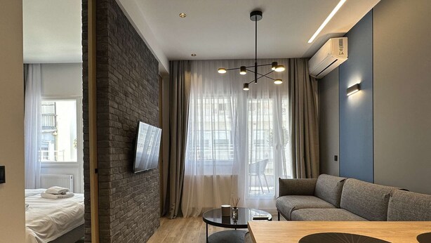Una habitación de hotel moderna con cama, sofá, mesa de comedor, televisor montado en la pared y un ventanal con cortinas.