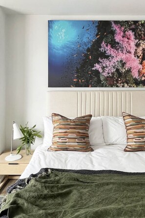 Een slaapkamer met een bed, kussens, een nachtkastje, een lamp en een wanddecoratie.