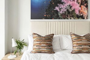 A bed with white bedding and striped pillows, a bedside lamp, and a plant on a wooden nightstand.