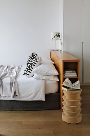 A neatly made bed with white linens and patterned pillows, a bedside table with a lamp and a glass of water, and a wooden bookshelf with books. - Unique Studio Apartment - Number 4704 (Cape Town)