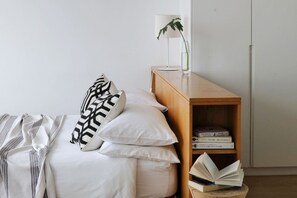 A neatly made bed with white bedding and patterned pillows, a bedside table with a lamp and a book, and a wooden bookshelf with books. - Unique Studio Apartment - Number 4704 (Cape Town)