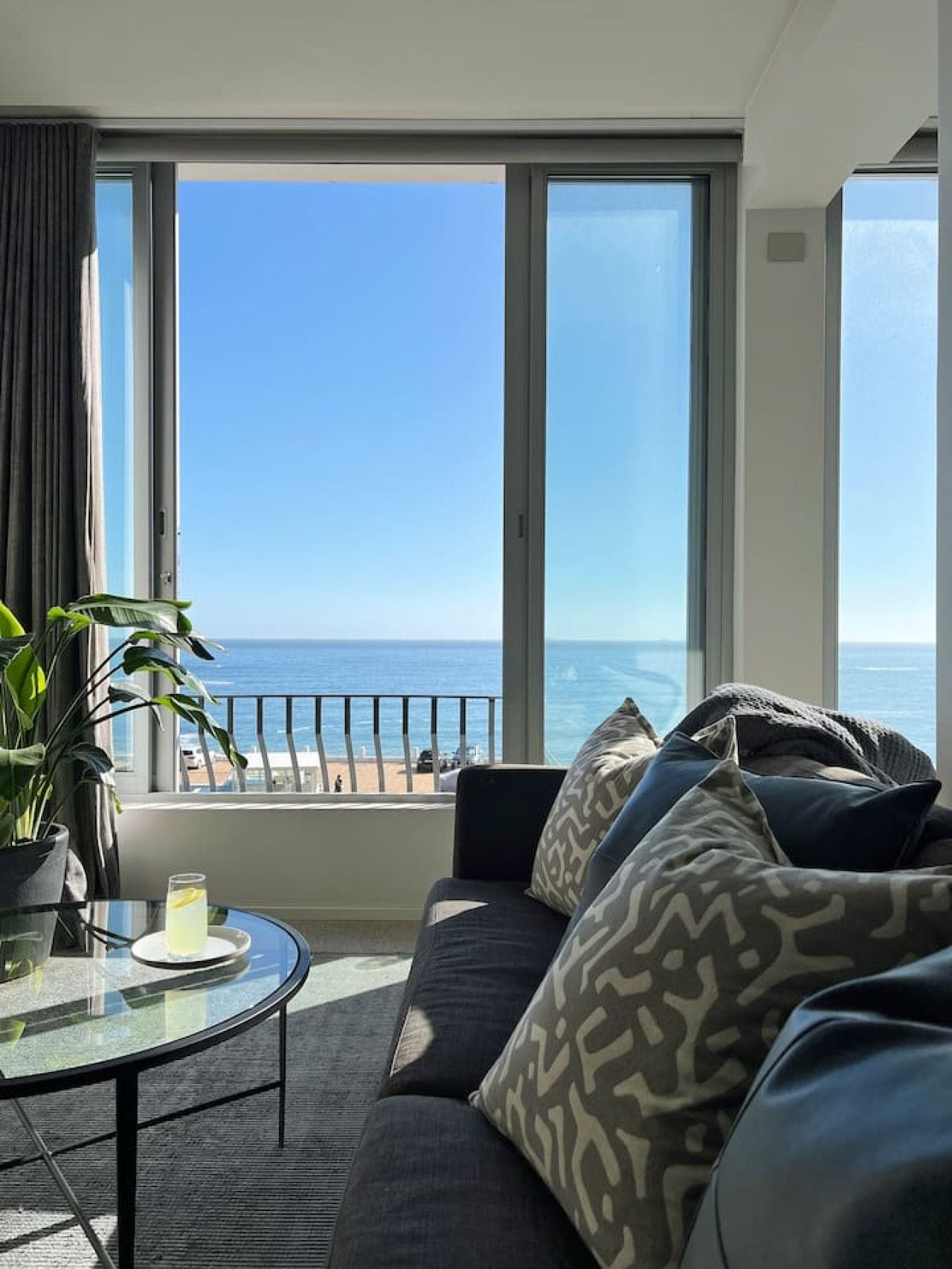 A modern living room with a large window overlooking a beach, a glass table with a drink, and a dark-colored sofa with patterned cushions.