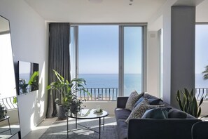 A modern living room with a balcony overlooking the sea, a sofa, a coffee table, and potted plants.