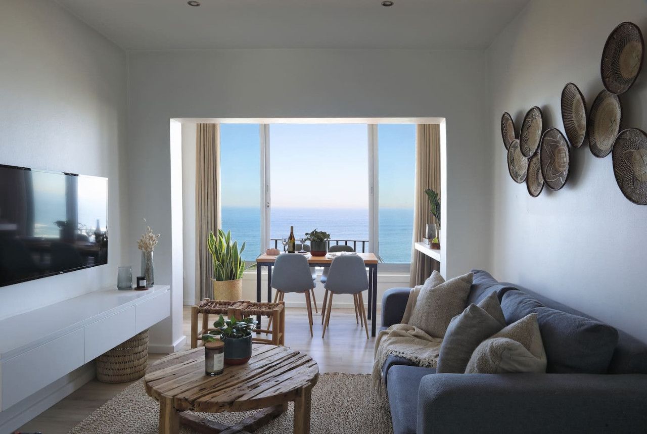 Una sala de estar moderna con un televisor grande, una mesa de centro de madera, un sofá y un comedor con vista al mar.