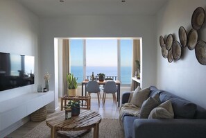 A modern living room with a flat-screen TV, a wooden coffee table, a grey sofa, and a dining area with a view of the sea. - Cozy Studio Apartment - Number 4705 (Cape Town)