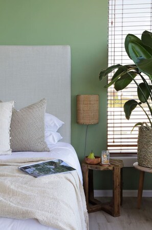 A bedroom with a bed, pillows, a bedside lamp, a plant, and a small table with a bowl and a glass.