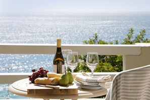 Ein Tisch ist reichhaltig mit Wein, Gläsern, Trauben, Brot und Birnen gedeckt, auf einem Balkon mit Blick aufs Meer.