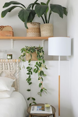 Ein Schlafzimmer mit einem Bett, einem Nachttisch mit einer Zeitschrift, einer Lampe und zwei Regalen mit Pflanzen und einem Korb.