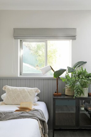 A bedroom with a bed, a bedside lamp, a potted plant, and a window with a view of greenery.