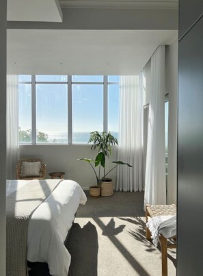 Ein Schlafzimmer mit großem Fenster, einem Bett, einem Sessel, einem kleinen Tisch und einer Zimmerpflanze.