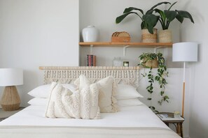 A neatly arranged bedroom with a bed, pillows, a bedside lamp, a small table, a shelf with books and a vase, and a hanging plant.