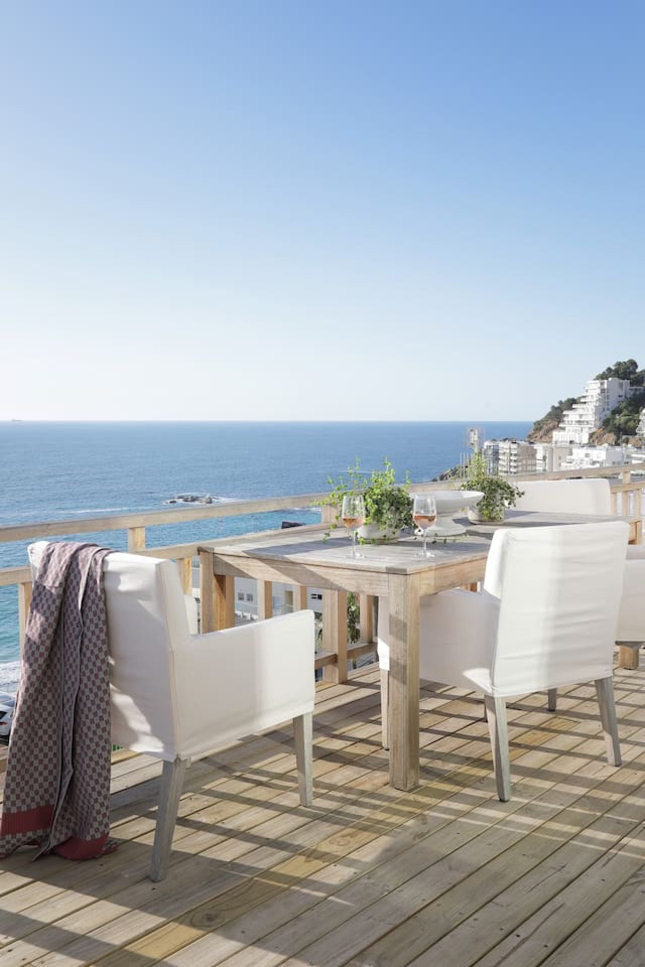An outdoor dining area with a view of the sea, wooden flooring, and white chairs.