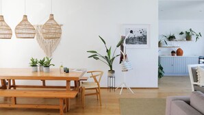 A modern living space with a wooden dining table, chairs, hanging woven light fixtures, a potted plant, a framed picture, and a kitchen area with cabinets and plants. - Beach House - Number 49 (Cape Town)