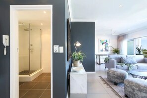 A modern interior with a glass-enclosed shower, a seating area with a sofa and ottoman, and a small table with a lamp. - Contemporary Clifton Apartment - Number 5104 (Cape Town)