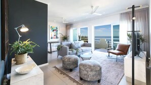 Ein modernes Wohnzimmer mit einer Couch, Poufs und einem Couchtisch. Ein Balkon bietet Blick auf den Himmel.