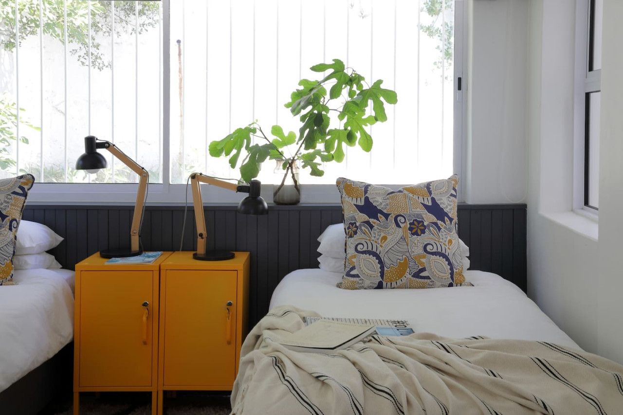A bedroom with a bed, a nightstand, a lamp, a plant, and a window with blinds.