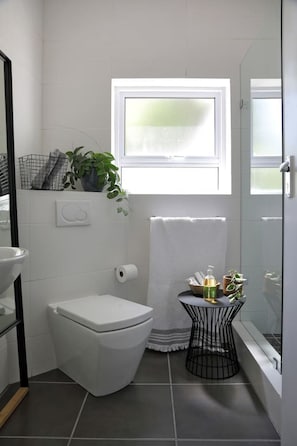 A compact bathroom with a toilet, a small round table with toiletries, a window with a view, and a towel rack. - Contemporary Clifton Apartment - Number 5104 (Cape Town)