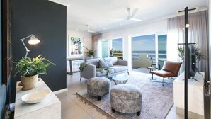 Ein modernes Wohnzimmer mit einem grauen Sofa, Poufs und einem Couchtisch. Ein großes Fenster bietet Blick auf das Meer.