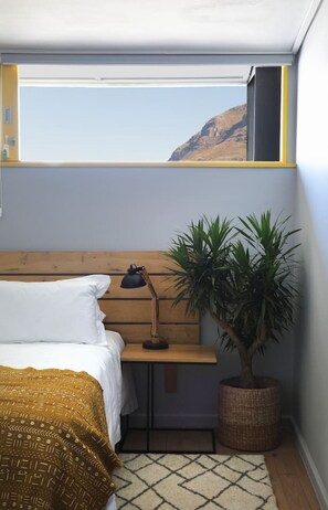 A bedroom with a bed, a desk, a lamp, a potted plant, and a view of a mountain.