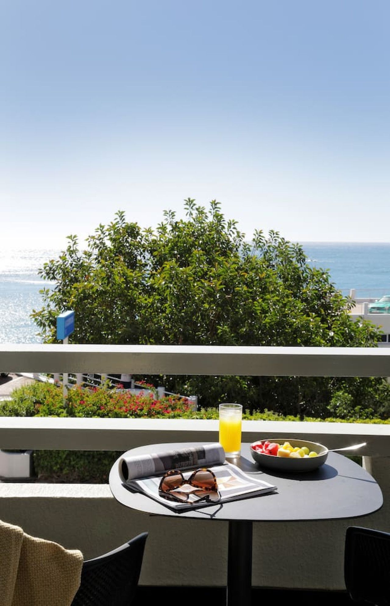 A balcony with a table, a bowl of fruit, a glass of orange juice, and a pair of sunglasses.