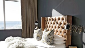 Un dormitorio con cabecera capitonada, lámpara de noche, almohadas estampadas, manta suave y vistas de la ciudad a través de una ventana.